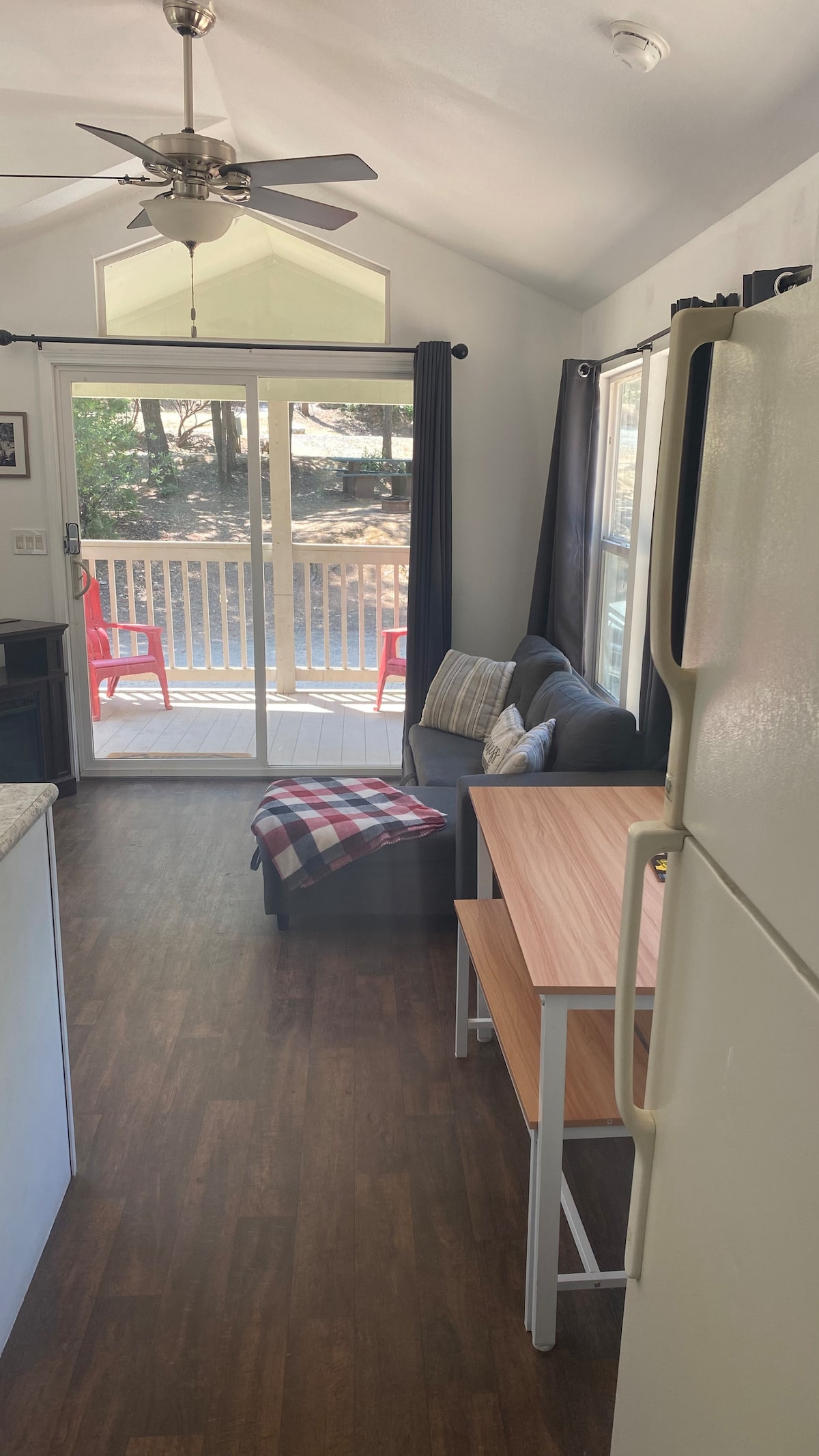 A comfortable living area features a grey couch with decorative pillows and a wooden dining table. Large glass doors provide access to a deck, while natural light brightens the space. A ceiling fan adds to the cozy atmosphere, complemented by dark curtains.