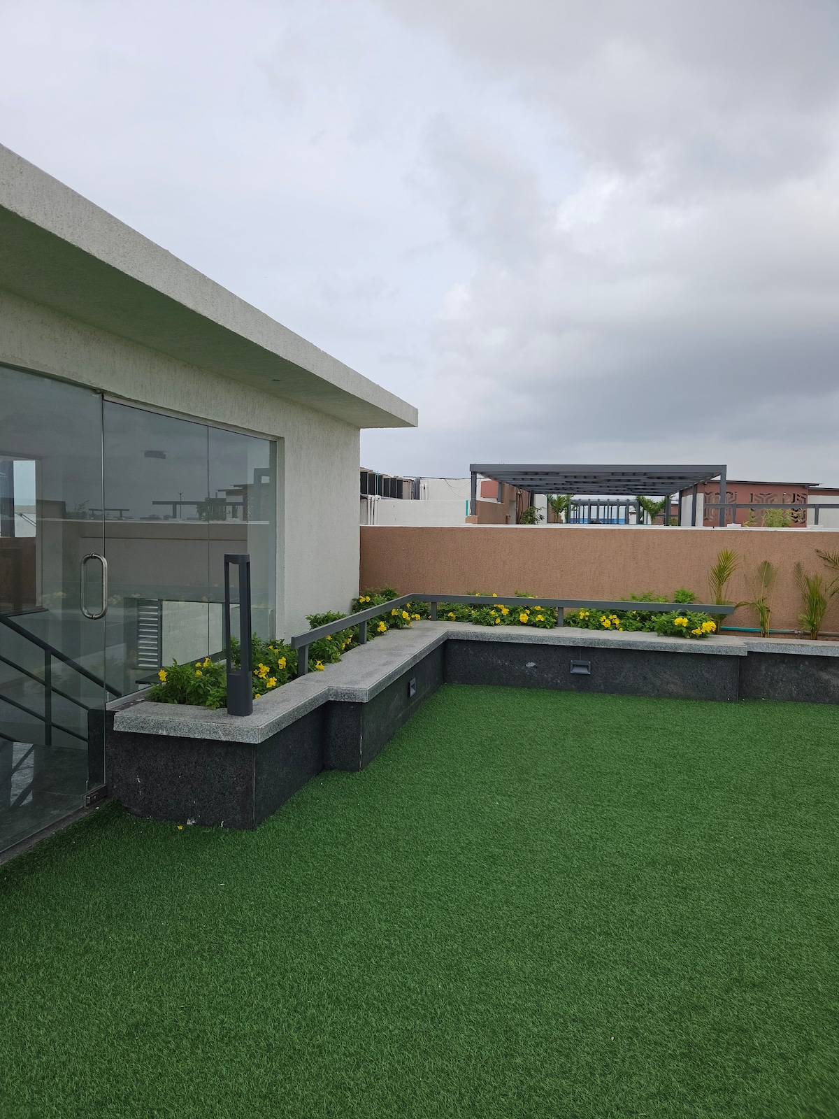 A spacious rooftop area is depicted, featuring artificial grass and a stone bench adorned with vibrant yellow flowers. Large glass doors lead into the interior, providing a seamless transition from inside to outside.
