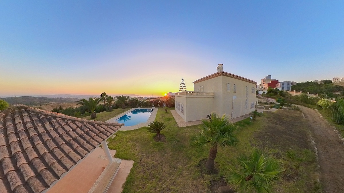 A panoramic view captures a villa alongside a shimmering private pool, with lush greenery and palm trees adorning the landscape. The sun sets on the horizon, casting warm hues over the scene, while a neighboring structure is visible to the right.