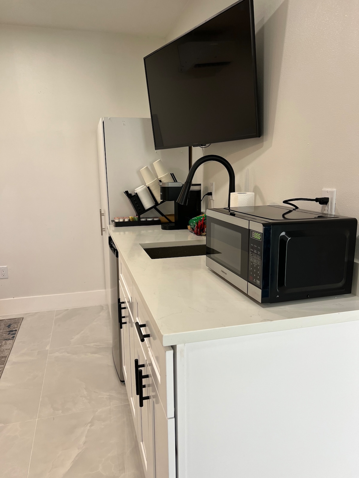 A modern kitchen area features white cabinetry with sleek black handles. A microwave and a coffee maker are positioned on the countertop, accompanied by a small sink underneath a mounted television. Light tiles enhance the bright and airy atmosphere.