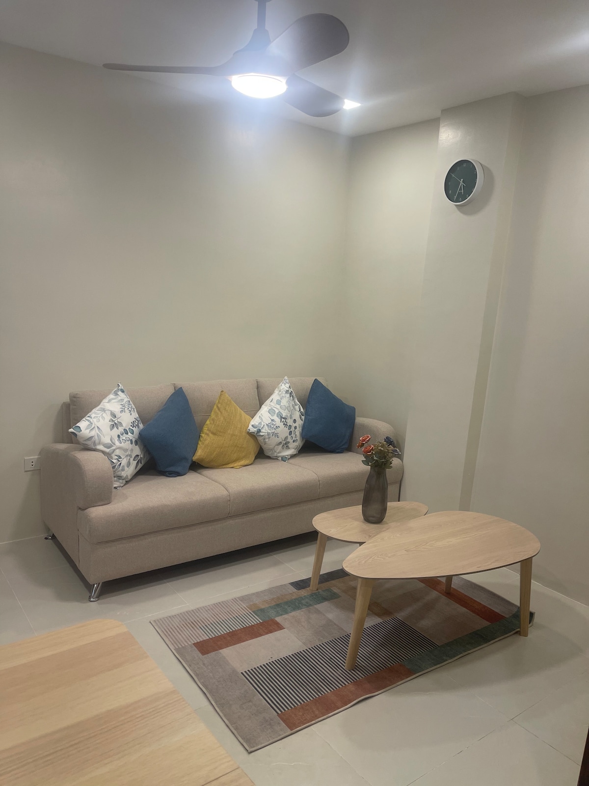 A cozy seating area is presented with a light-colored sofa adorned with colorful cushions. A wooden coffee table is positioned in front of the sofa, accompanied by a small vase with flowers. A clock is mounted on the wall, and a patterned area rug enhances the space.