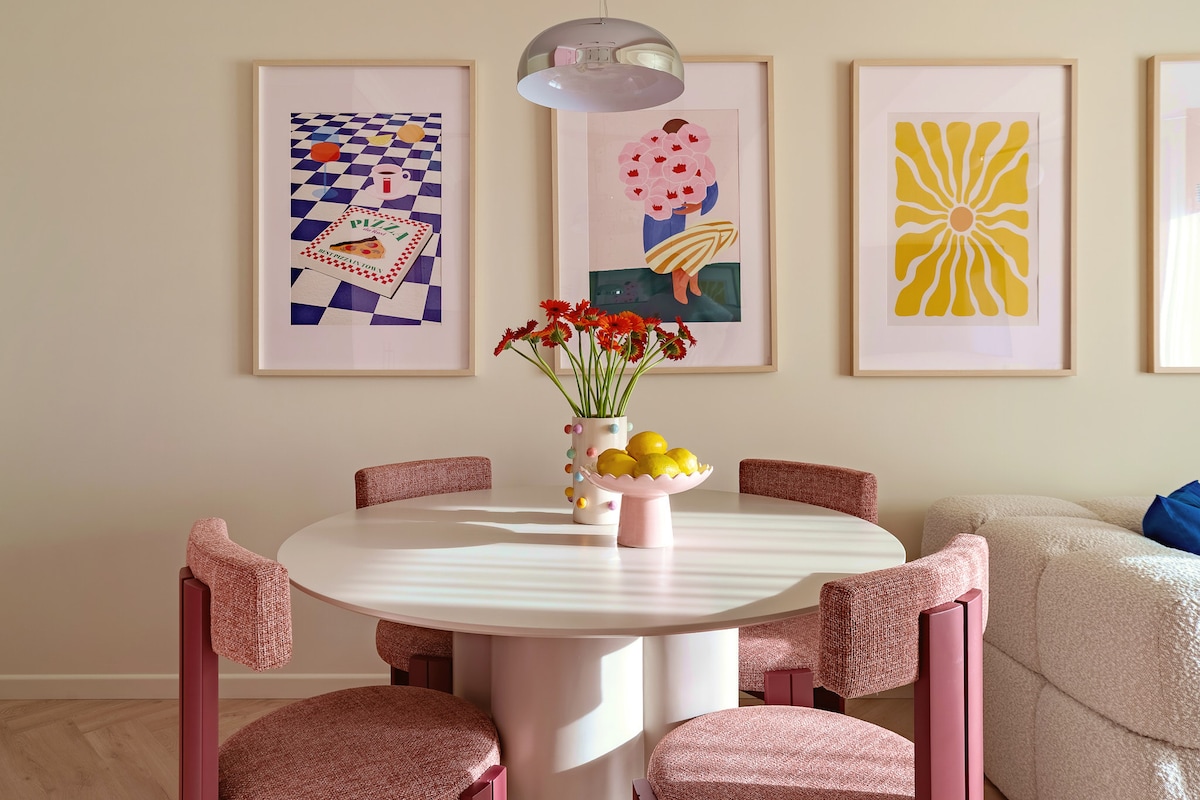 A round dining table is centered in a brightly lit area, surrounded by four chairs with soft, upholstered seating. Colorful art pieces adorn the walls, while a vase of flowers and a bowl of lemons are placed on the table, adding a touch of warmth to the space.