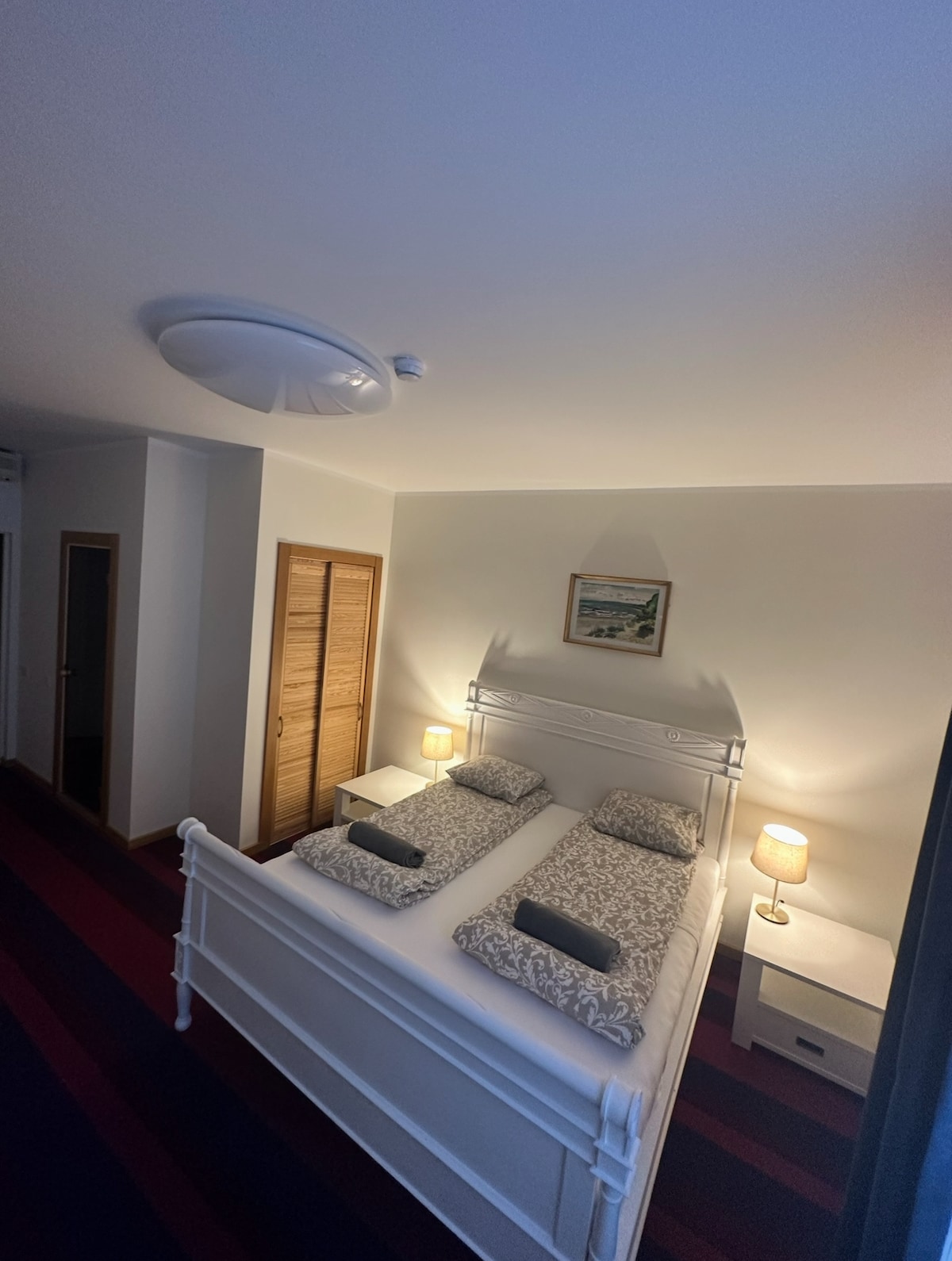 A serene bedroom is highlighted by a spacious white bed with two neatly arranged linens and contrasting gray pillows. Warm light from bedside lamps enhances the calming ambiance. An art piece featuring a landscape hangs above the bed, complementing the neutral wall color. A wooden wardrobe is visible.