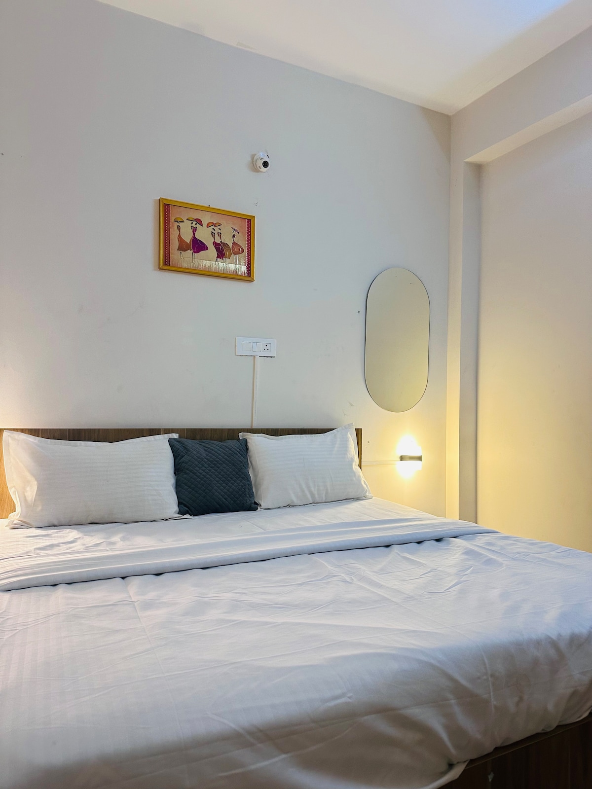 A cozy sleeping area features a spacious bed adorned with crisp white linens and two neatly arranged pillows. An artistic wall piece adds character above the bed, while a soft light source enhances the room's tranquility. A round mirror is mounted on the wall, complementing the overall decor.