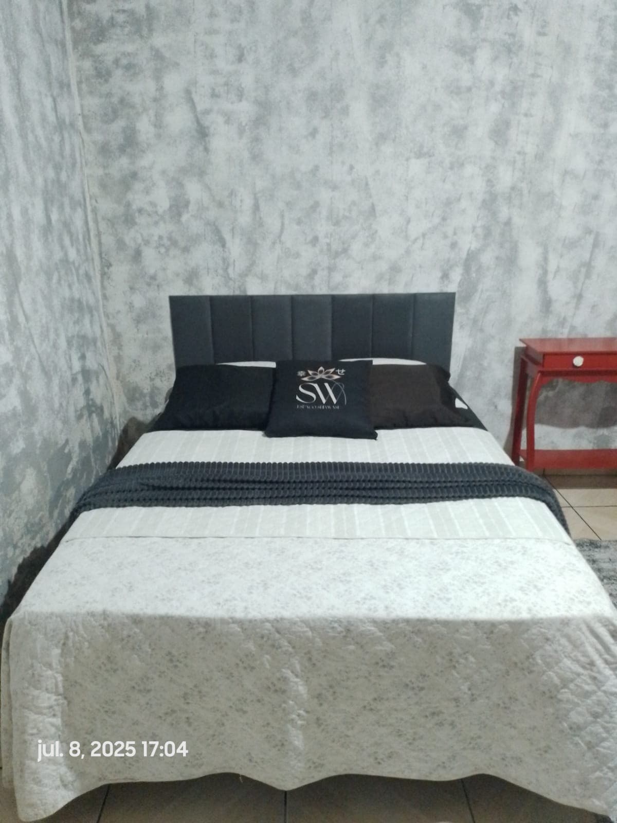 A comfortable double bed is positioned against a textured gray wall. The bed features a soft, neutral-toned coverlet with a contrasting dark throw at the top. A decorative pillow rests in the center, while a small red nightstand stands nearby.