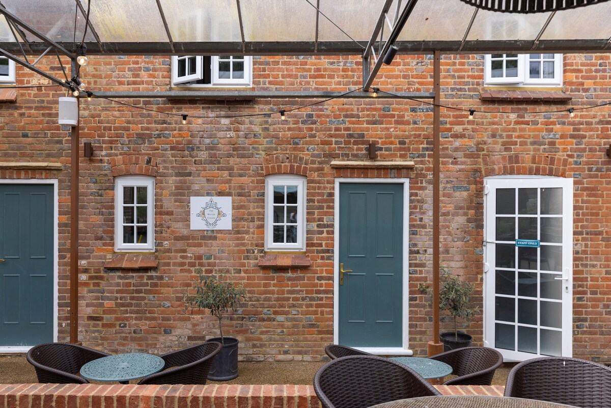 The exterior of the building features a rustic brick facade with three doors, two of which are traditional-style and one is framed by a glass panel. Outdoor seating is arranged with dark wicker furniture under a protective canopy.