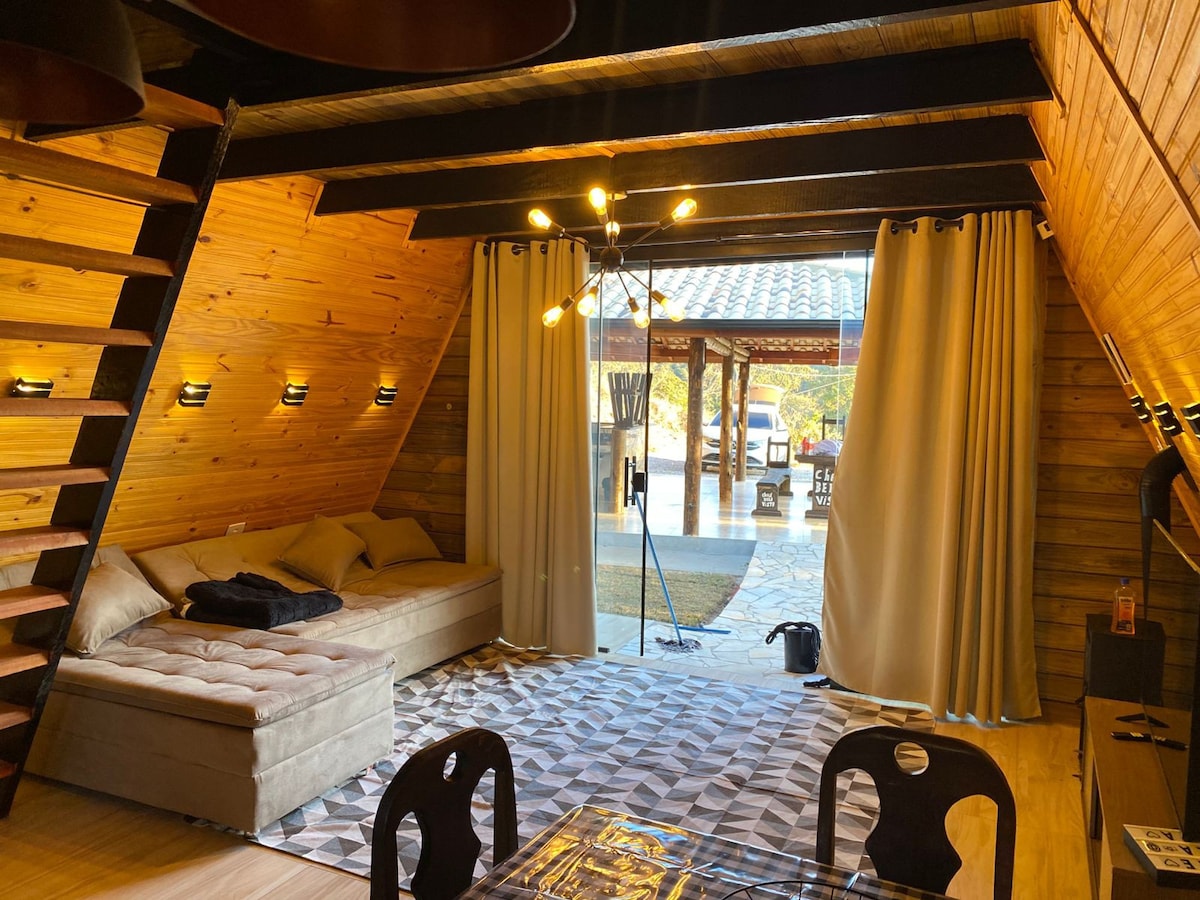 An inviting interior showcases wooden paneling and chic lighting fixtures. A spacious sofa is positioned against the wall, complemented by a textured area rug. Large, draped curtains frame views of an outdoor living space, allowing natural light to fill the room.