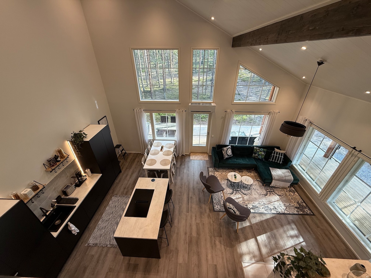 An overhead view of a spacious open-concept living area features large windows allowing natural light. The modern kitchen is equipped with a dark cabinetry, while a dining area with a circular table sits beside a living space with a dark green sofa and light-colored accent chairs.