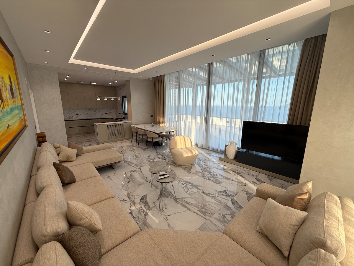 A spacious living area features a large sectional sofa, elegantly arranged around a central coffee table. Floor-to-ceiling windows provide panoramic views of the sea, while a modern dining table seats several guests. Light-colored marble floors enhance the airy ambiance of the space.