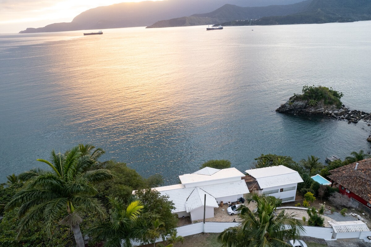 An aerial view captures a serene coastal landscape with a tranquil sea reflecting the warm hues of sunset. A charming white structure is nestled among green palm trees, with rocky outcrops and vessels visible in the distance, conveying a peaceful atmosphere.