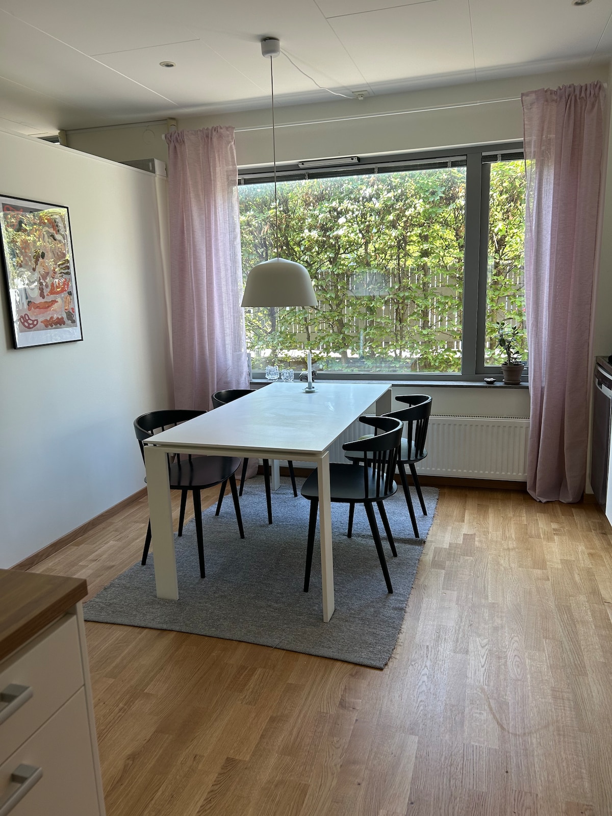 A dining area features a white table surrounded by four black chairs on a light rug. Natural light enters through a large window adorned with sheer curtains, offering views of greenery outside. The space is complemented by a simple pendant light above the table.