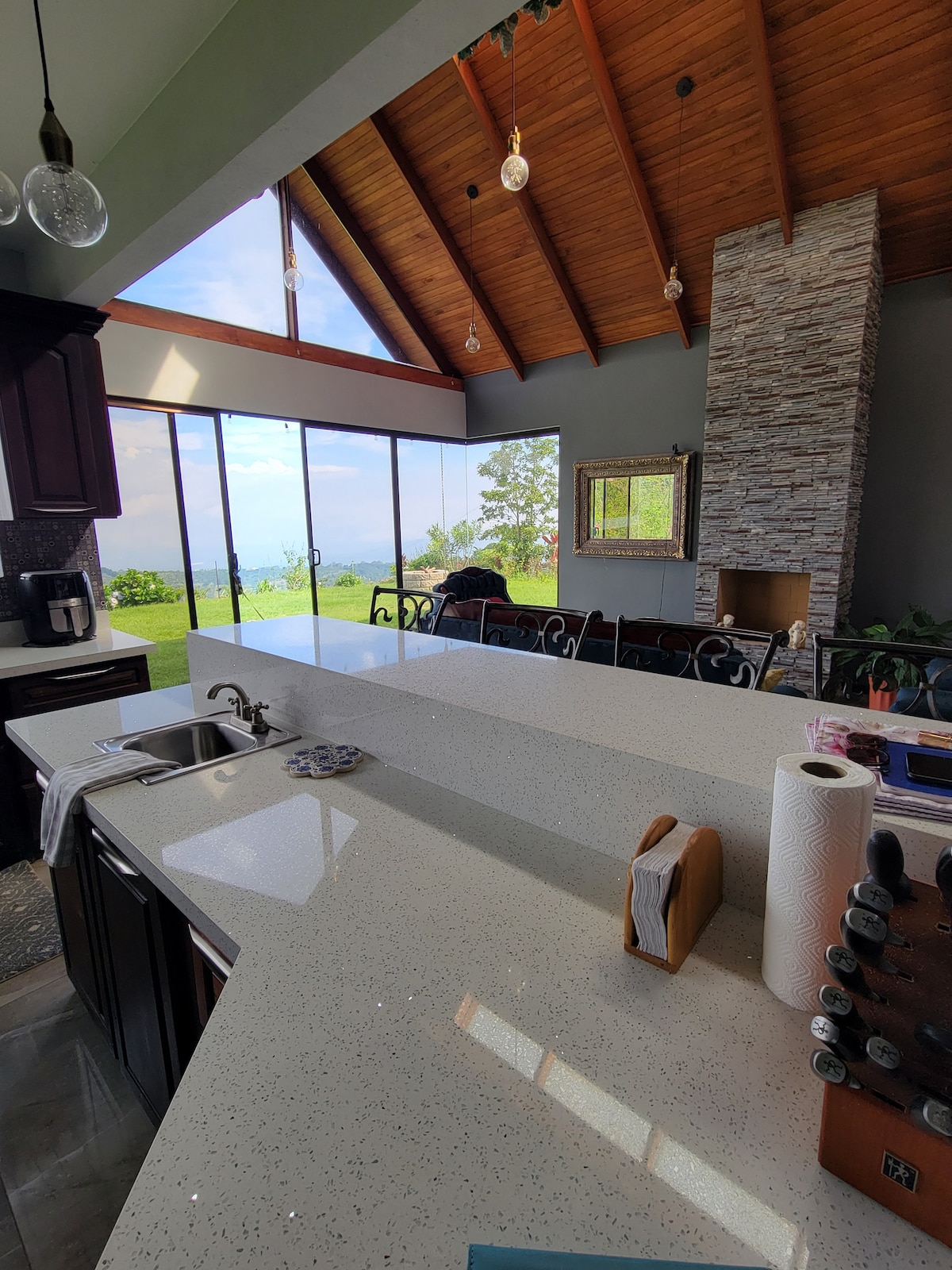 A modern kitchen features an expansive quartz countertop with a sleek sink, overlooking large glass windows that provide views of the greenery outside. The high wooden ceiling adds warmth, complemented by a stone fireplace, creating a bright and airy atmosphere.