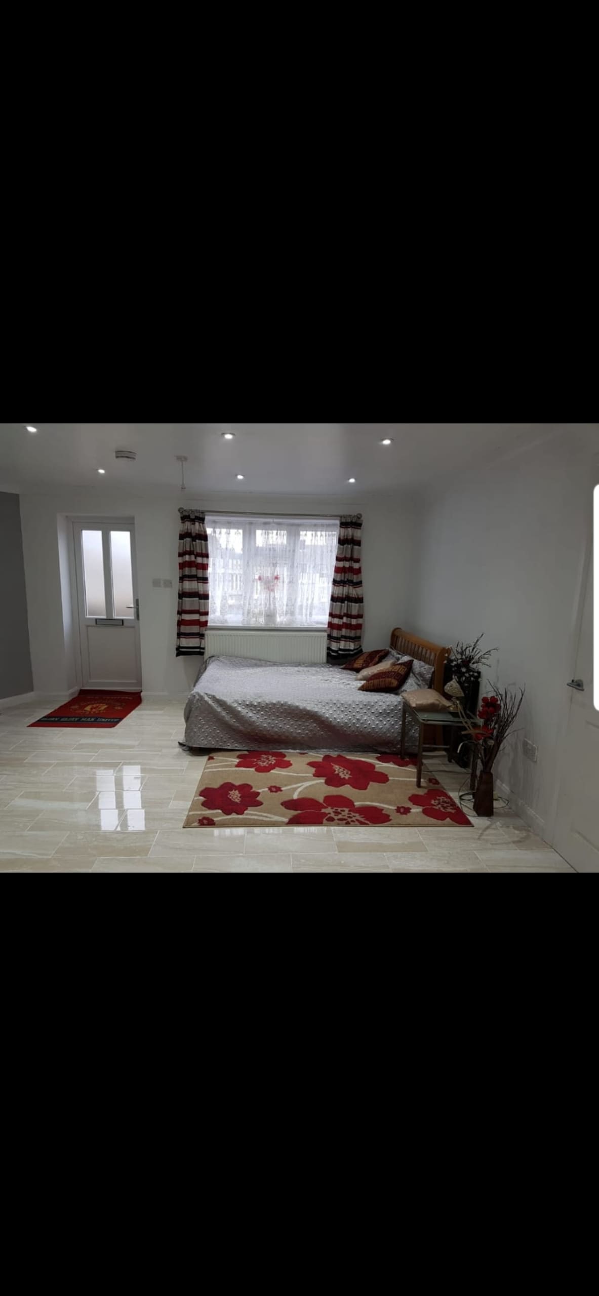 A spacious sleeping area features a double bed with a patterned coverlet, accompanied by a decorative striped curtain over the window. Accents of red and beige are seen in the rugs. Natural light enters from the window, illuminating the room's light-colored tiles.
