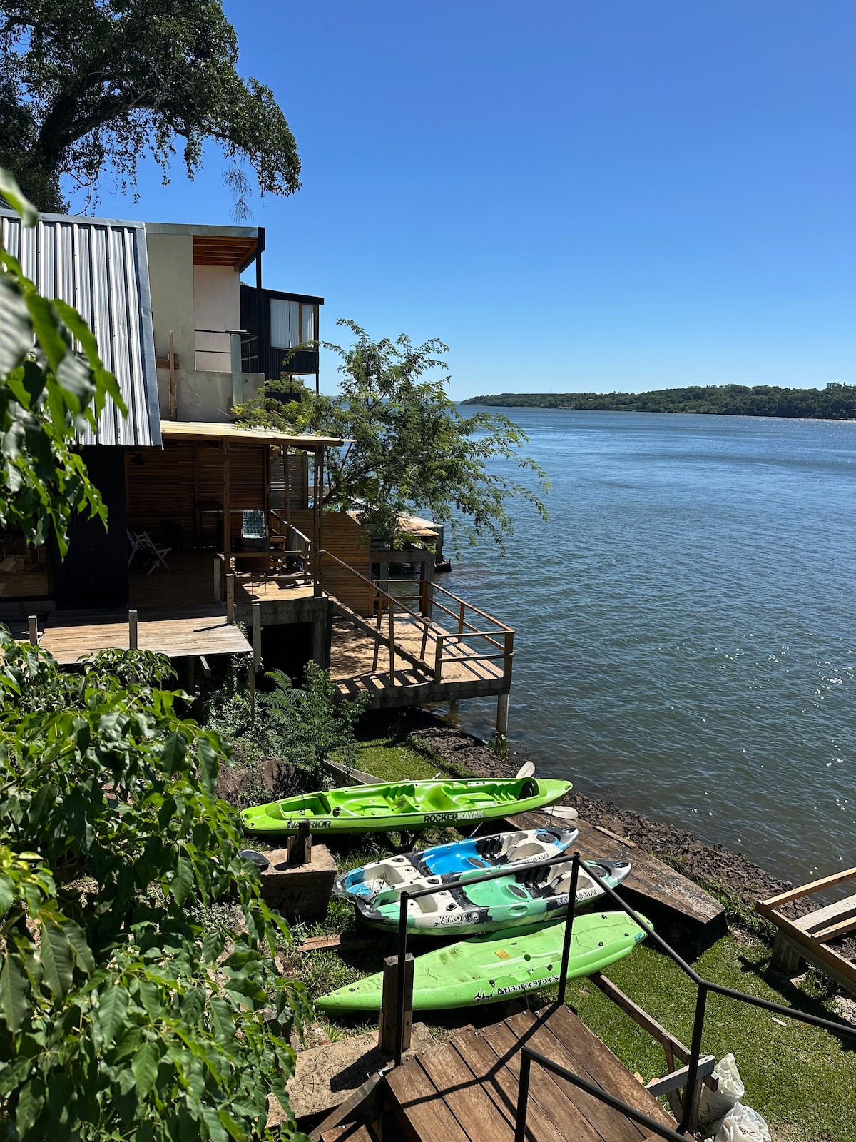A view of the riverbank featuring kayaks in vibrant green colors positioned along the water's edge. A wooden deck extends from a rustic building, providing access to the river. Lush greenery surrounds the area, enhancing the natural setting along the Paraná River.