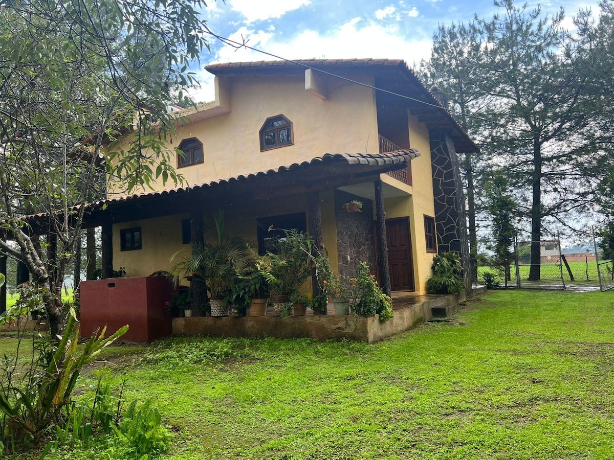 A two-story house is positioned amidst lush greenery, featuring a tiled roof and decorative stone accents. Surrounding the home, various plants and trees create a natural setting. The entrance is marked by a wooden door and a small covered porch.