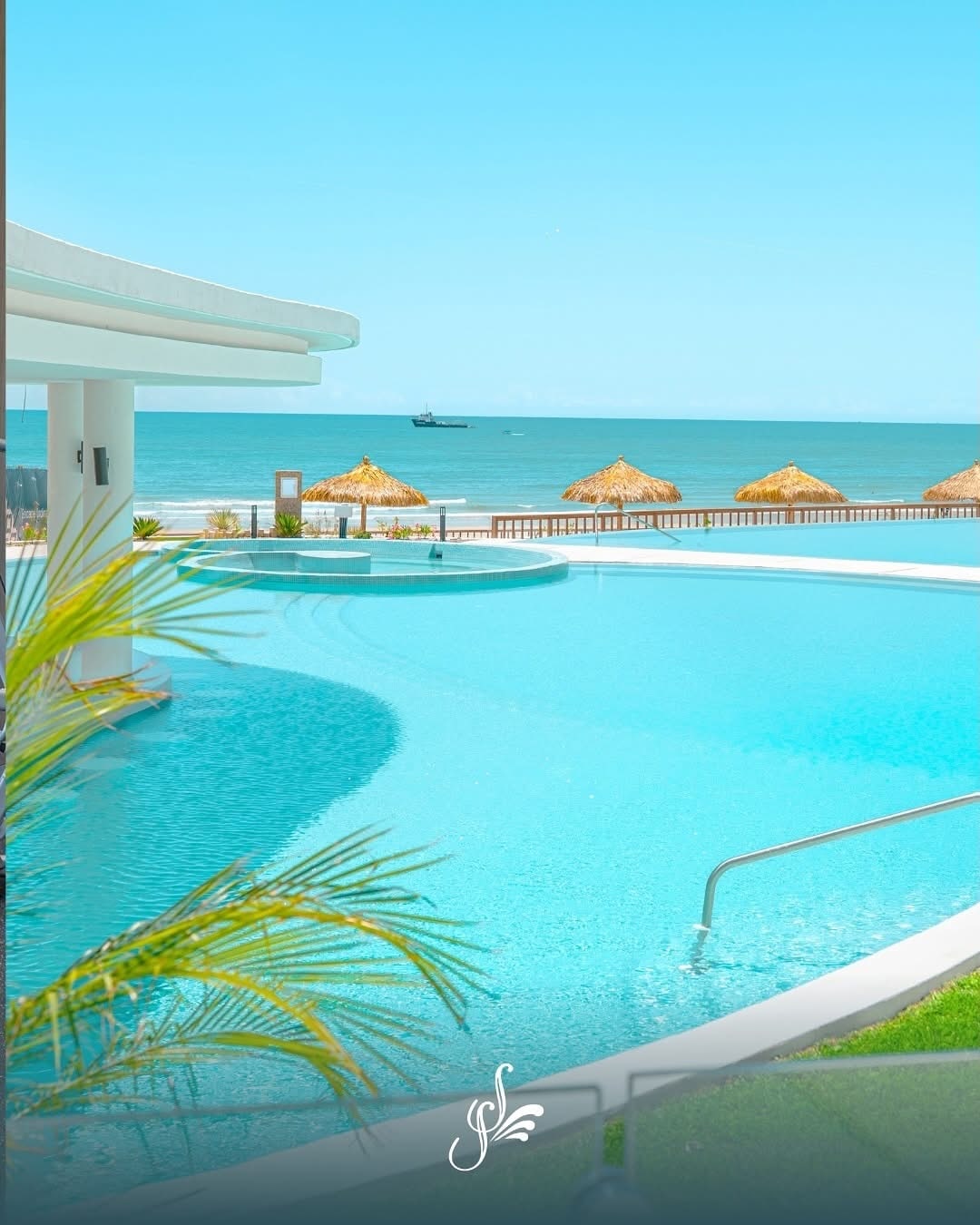 A large swimming pool is set against a backdrop of the ocean, with thatched umbrellas positioned along the shore. The pool features gentle curves and a bright blue hue, reflecting the clear sky above. Lush greenery borders the area, enhancing the serene environment.