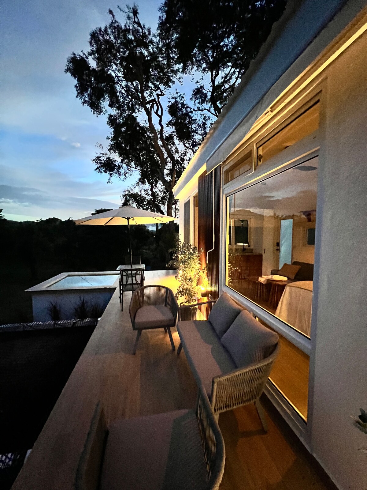 A serene outdoor seating area features two comfortable chairs and a small side table, with soft lighting highlighting the space. A large umbrella provides shade, while a glimpse of a tranquil pool is visible in the background, surrounded by greenery under a twilight sky.