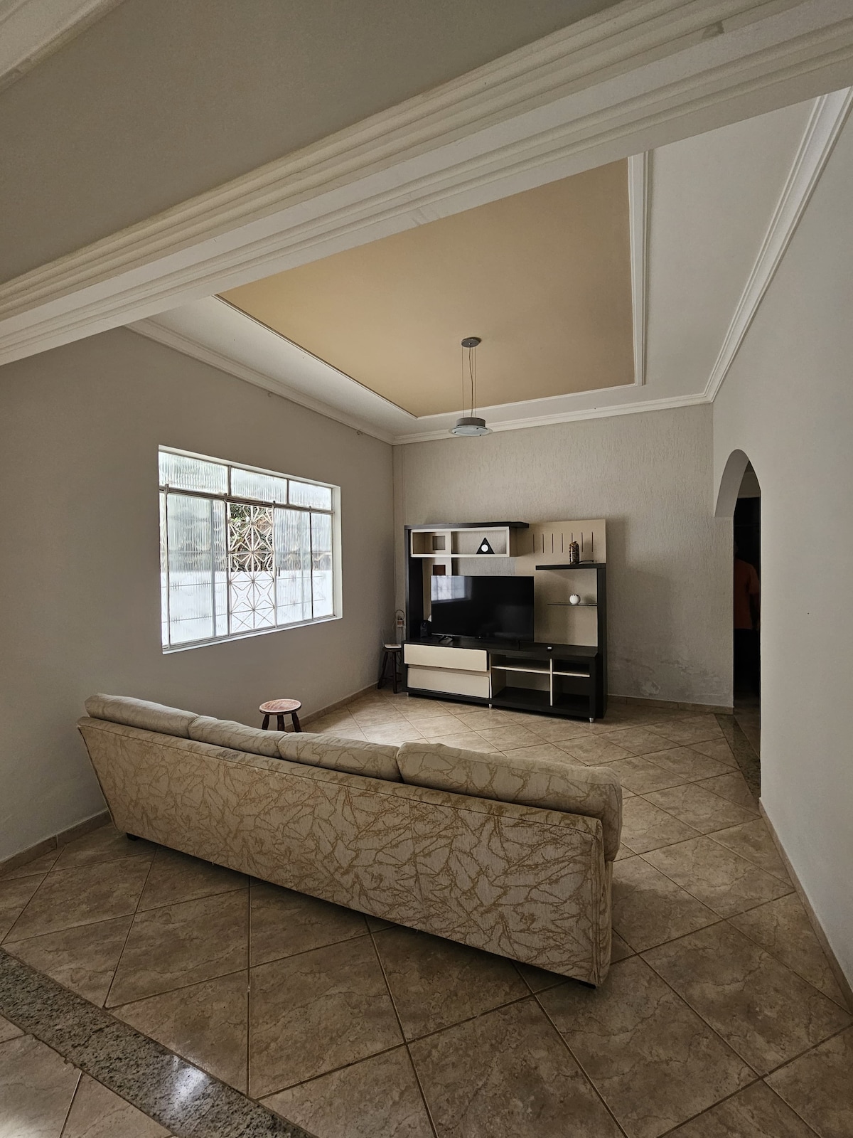 A living area is characterized by a comfortable sofa facing a mounted television. A light-colored shelf displays decorative items, and a small table is positioned nearby. Natural light enters through a large window, highlighting the textured walls and tiled floor.
