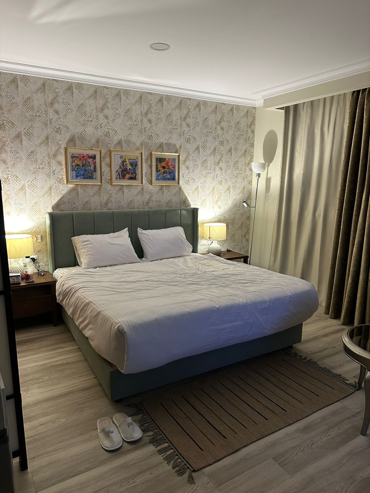 A tranquil bedroom features a spacious bed with white linens and two plush pillows. Warm lighting from bedside lamps provides a soft glow. The walls are decorated with framed artwork, and a neutral rug adds comfort underfoot. Thick curtains frame a window, enhancing the relaxing ambiance.
