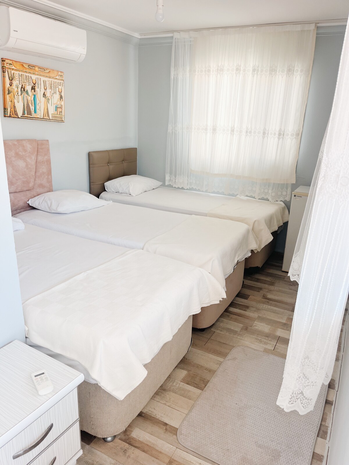 A bright sleeping area contains two beds with soft white linens, arranged side-by-side. A window is adorned with a delicate lace curtain, allowing natural light to filter in. A light wooden floor adds warmth to the space, complemented by a small bedside table.