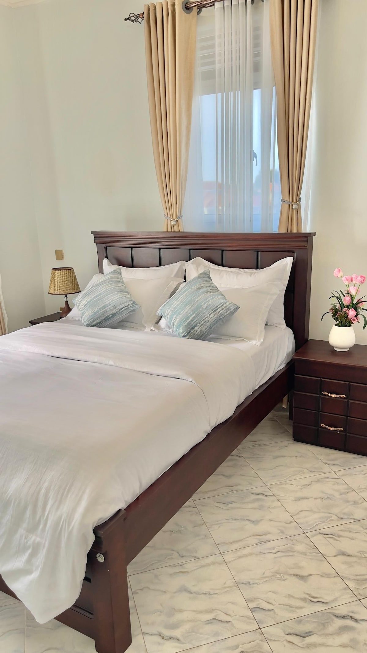 A comfortable sleeping area is featured, including a spacious bed with light bedding accented by blue decorative pillows. Natural light streams through a large window dressed with sheer curtains, while a bedside table with a lamp and a small vase of flowers adds subtle decor.