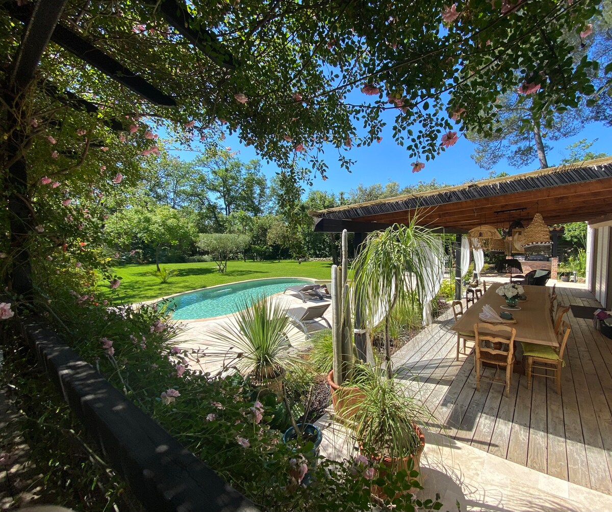 The outdoor area features a spacious deck lined with greenery and flowers. A private pool reflects sunlight, surrounded by lush lawns. An outdoor dining space equipped with a wooden table and chairs is shaded by a pergola, inviting relaxation in a serene environment.