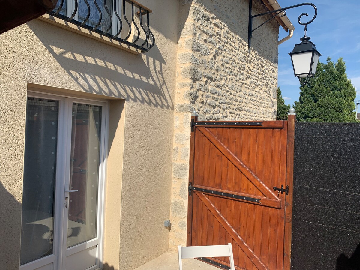 An exterior view of the lodging showcases a combination of smooth and textured walls. French doors are visible, allowing natural light to enter. A wooden gate adds a rustic touch, while a vintage lantern provides soft lighting. Surrounding greenery is also present in the background.