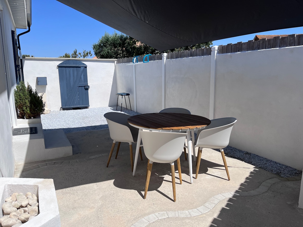 An outdoor dining area is presented with a round wooden table surrounded by four white chairs. A minimalistic building is visible in the background, along with a gravel pathway and a small storage shed. Sunlight illuminates the space, creating a bright atmosphere.