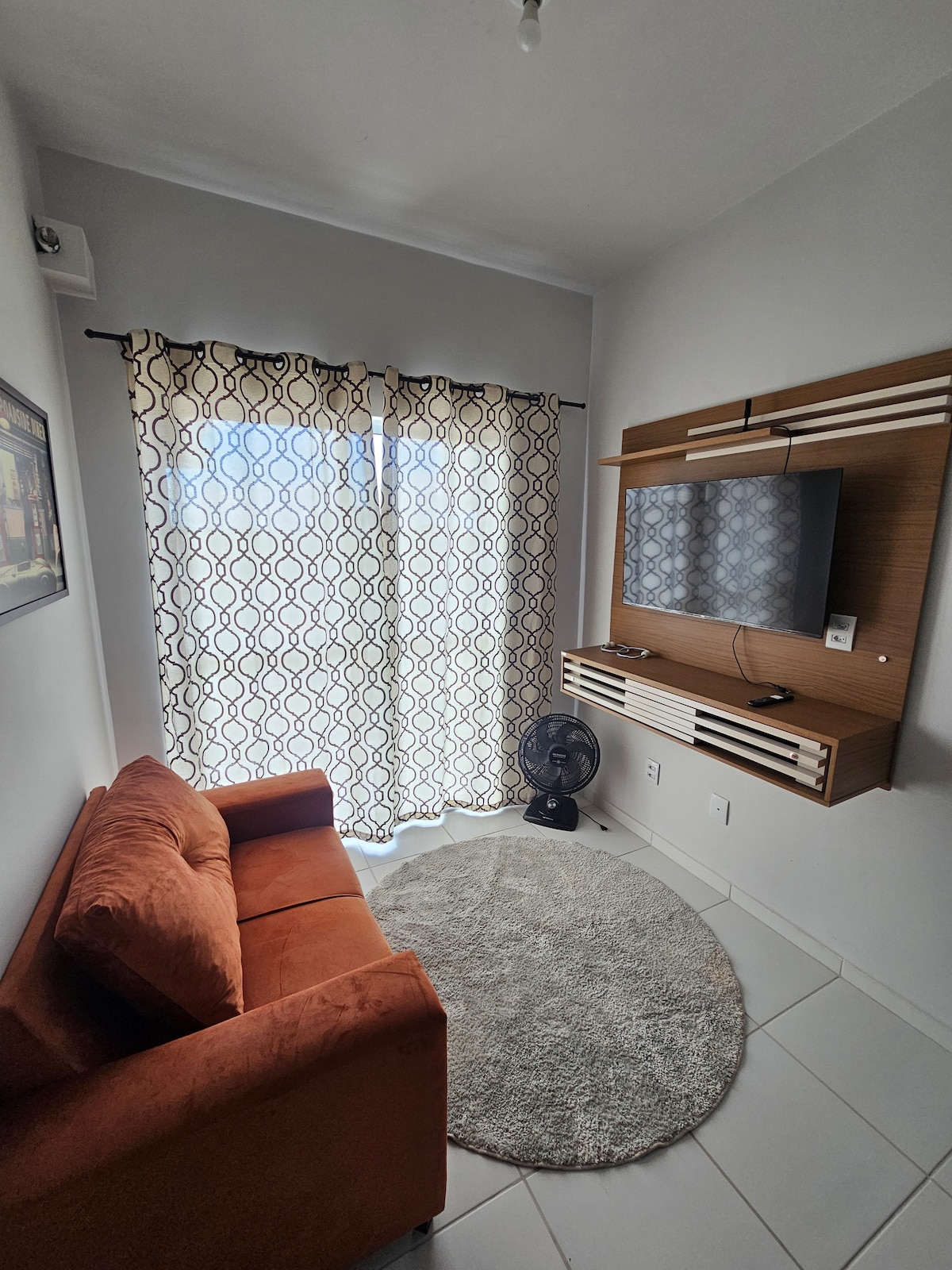 A living area features a cozy orange sofa and a circular rug on the tiled floor. Natural light enters through sheer curtains, which frame a large window. A modern wall-mounted television is positioned above a console, and a fan is placed on the floor for comfort.