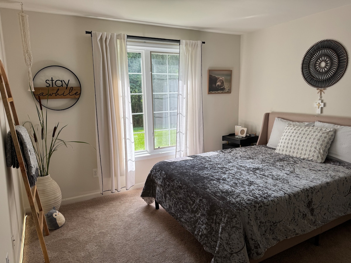 A serene bedroom features a spacious bed adorned with a textured gray blanket. Soft natural light filters through sheer white curtains, illuminating the room. A decorative wall sign and a stylish round wall piece add character, while a plant and an accent chair offer a cozy touch.