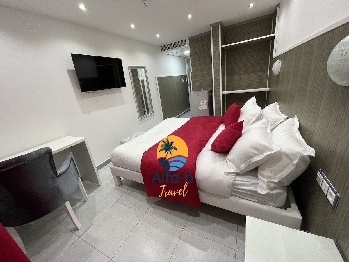 A well-appointed sleeping area features a spacious bed layered with white linens and red accent pillows. A compact desk and chair are positioned to provide workspace. Wall-mounted television and built-in shelving units are visible, enhancing the practical design of the room.