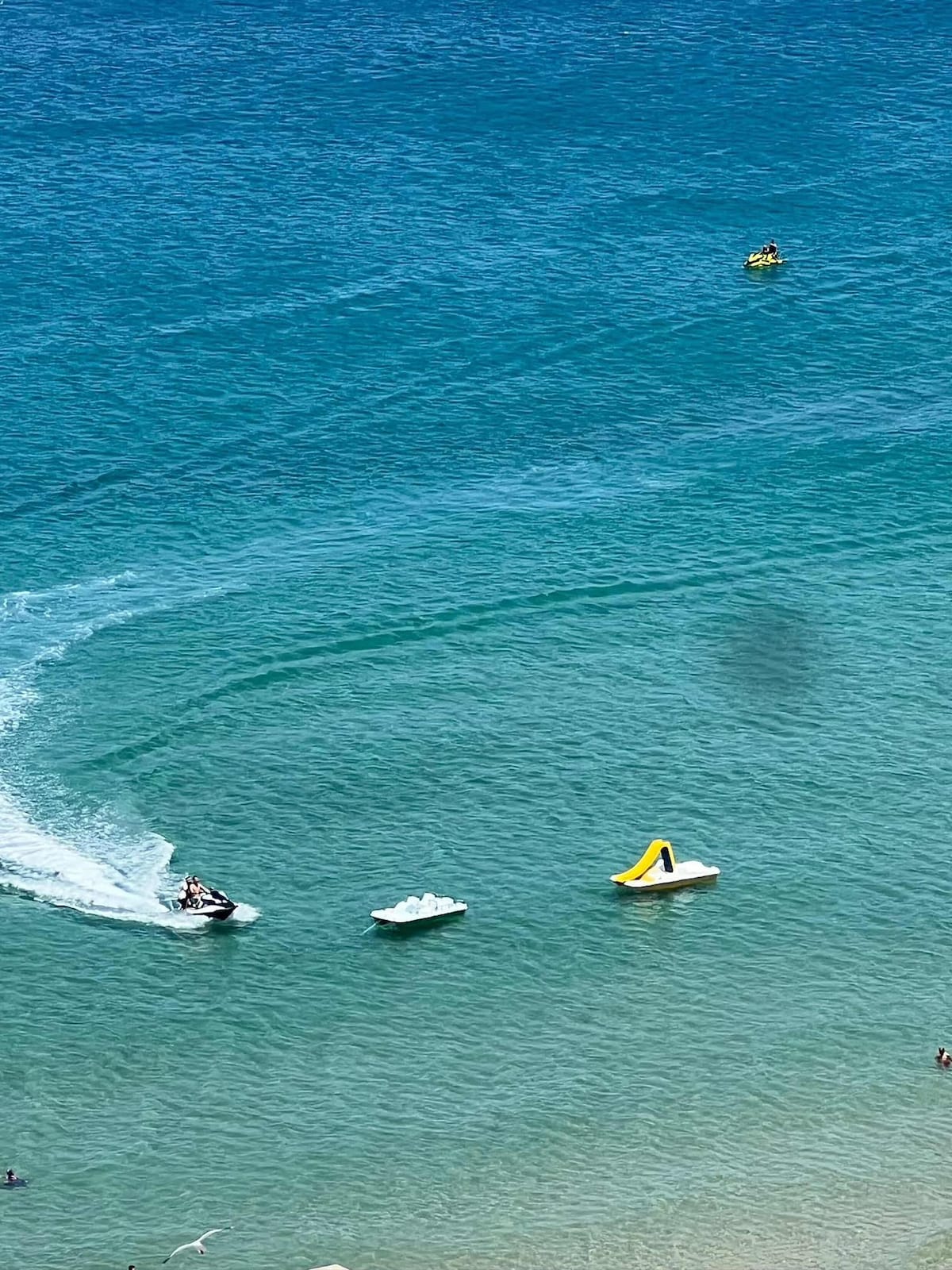 A clear blue sea is shown with gentle waves and a sandy shoreline. A jet ski is seen creating ripples in the water, while colorful inflatable equipment floats nearby. In the background, a distant group of people can be observed enjoying the water.