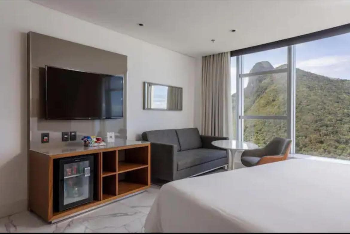 A well-appointed room features a spacious bed, a sleek television mounted on the wall, and a modern seating area. A window provides a view of the surrounding mountains, while a small table and chairs offer a space for relaxation. A minibar is located in the entertainment unit.