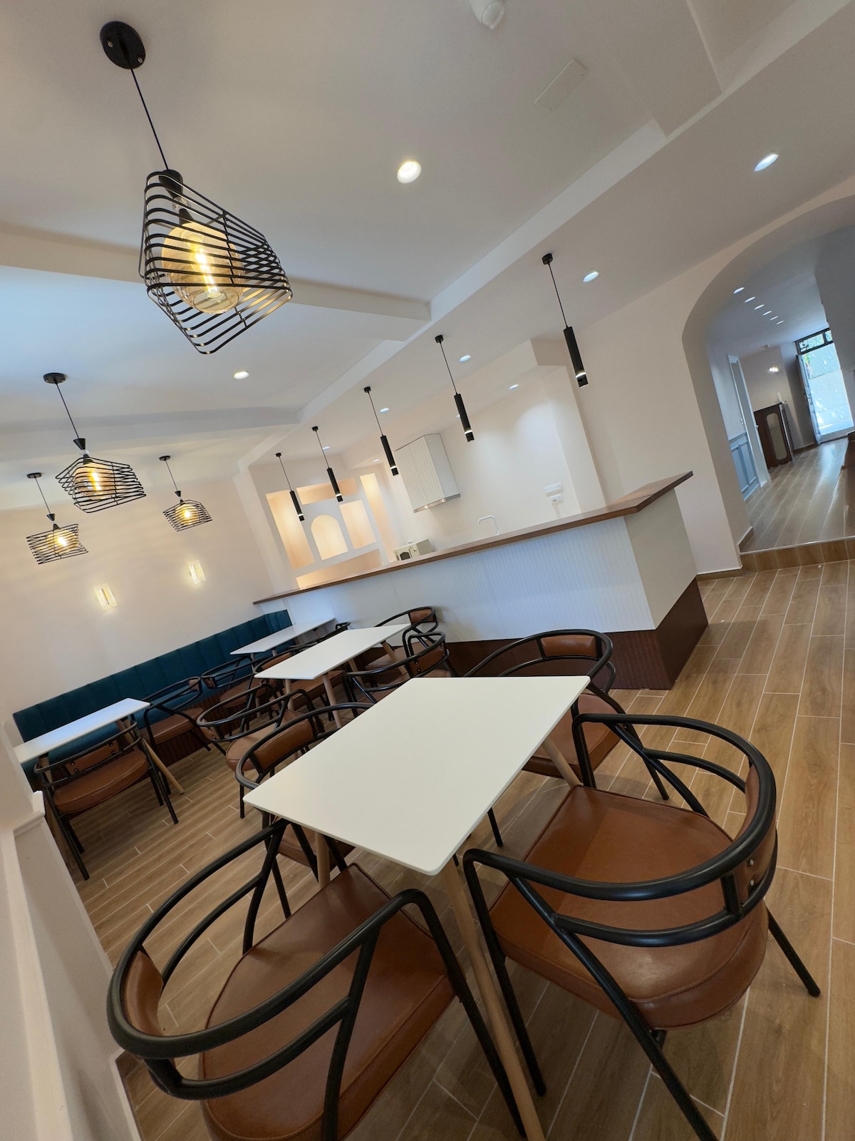 A modern dining area features light wood flooring and a bright, airy ambiance. Two tables are surrounded by brown metal-framed chairs. Pendant light fixtures provide illumination from above, while a bar area is visible in the background, enhancing the welcoming environment.