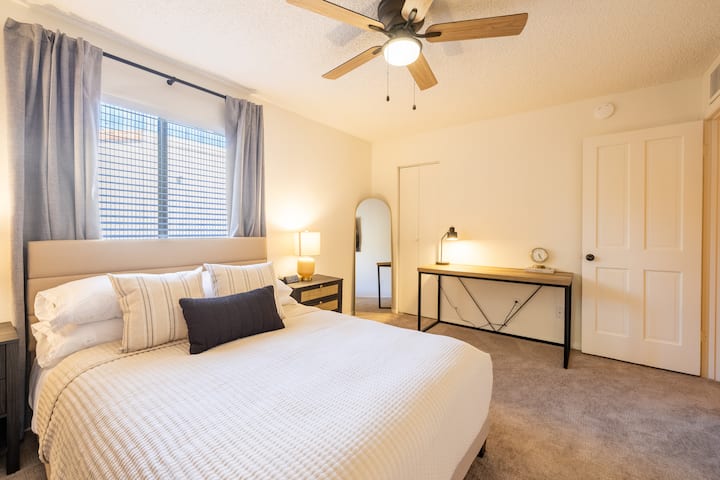 ห้องนอนใหญ่มีเตียงขนาดควีนไซส์ โต๊ะทำงาน สมาร์ททีวีขนาด 65” พอร์ตชาร์จบนโต๊ะข้างเตียง นาฬิกาปลุก และม่านบังแสง