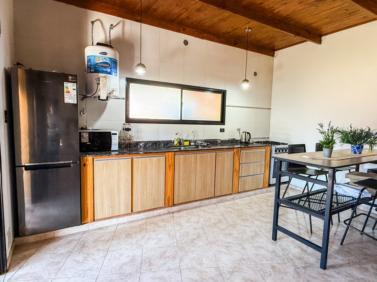 A spacious kitchen area is featured, equipped with modern appliances including a refrigerator and a stove. Cabinets and counter space are visibly organized, and a dining table with chairs is positioned nearby. Natural light shines through a large window, illuminating the warm wooden accents.