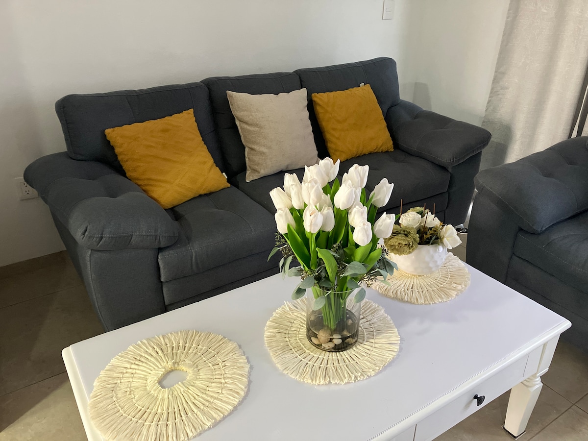 A comfortable seating area is presented with a dark gray sofa adorned with yellow and beige throw pillows. A white coffee table is centrally positioned, featuring a vase of white tulips and decorative elements placed on woven placemats.