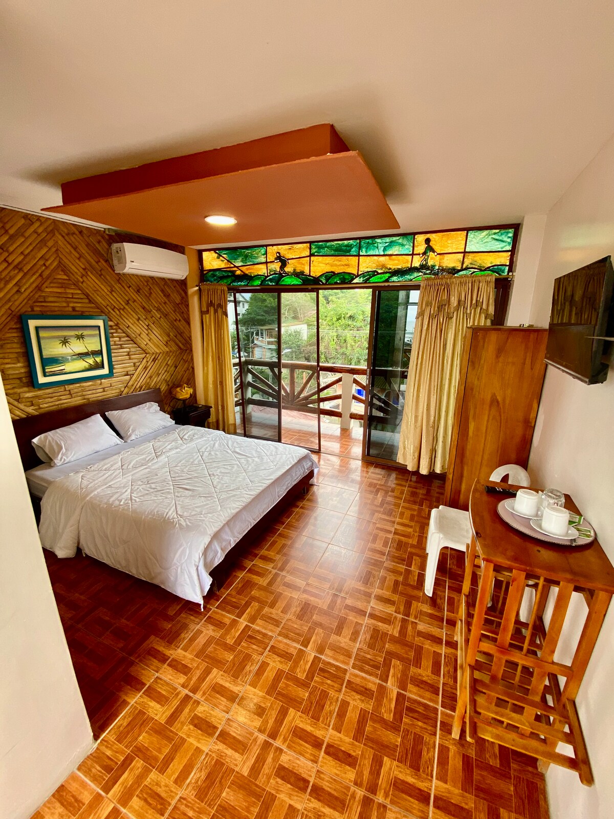 A well-lit room features a spacious bed with white linens against a backdrop of wooden accents. A small table with tea and coffee essentials is present, complemented by a white chair. Large glass doors open to a balcony, framed by golden curtains that softly diffuse natural light.