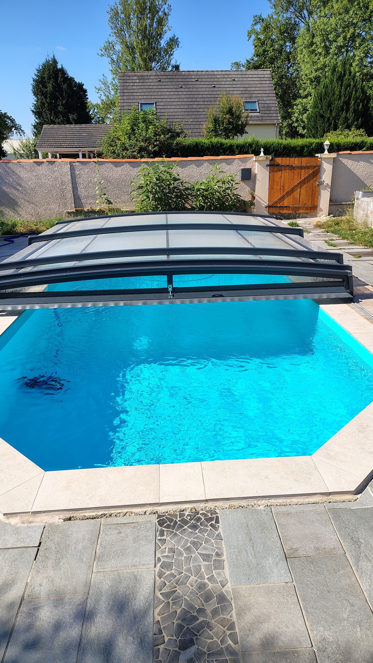 A well-maintained swimming pool is shown with clear blue water, surrounded by light-colored stone tiles. The pool area features a modern retractable cover and greenery is visible in the background, along with a wooden gate providing access to the property.