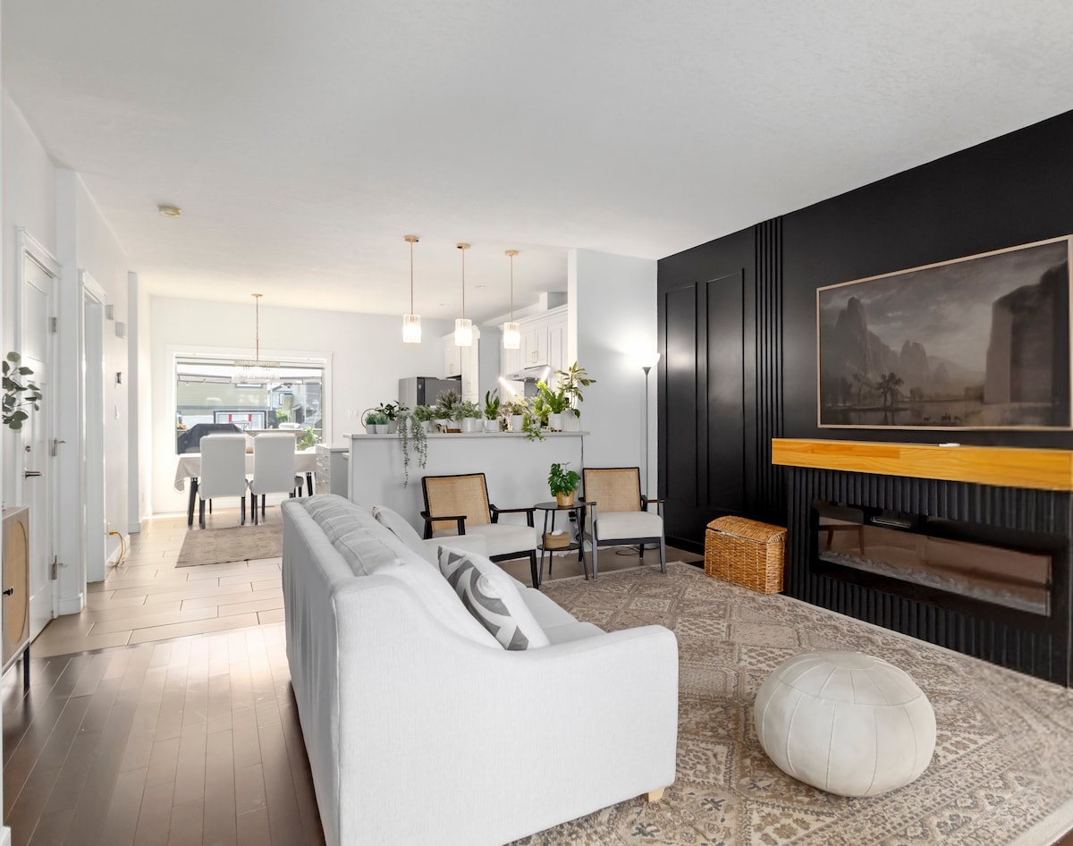 A spacious living area is highlighted by light-colored furnishings and a patterned rug. A cozy seating arrangement faces a modern entertainment unit, featuring a large artwork and a warm-hued accent wall. The open layout allows for visibility of the kitchen and outdoor area.