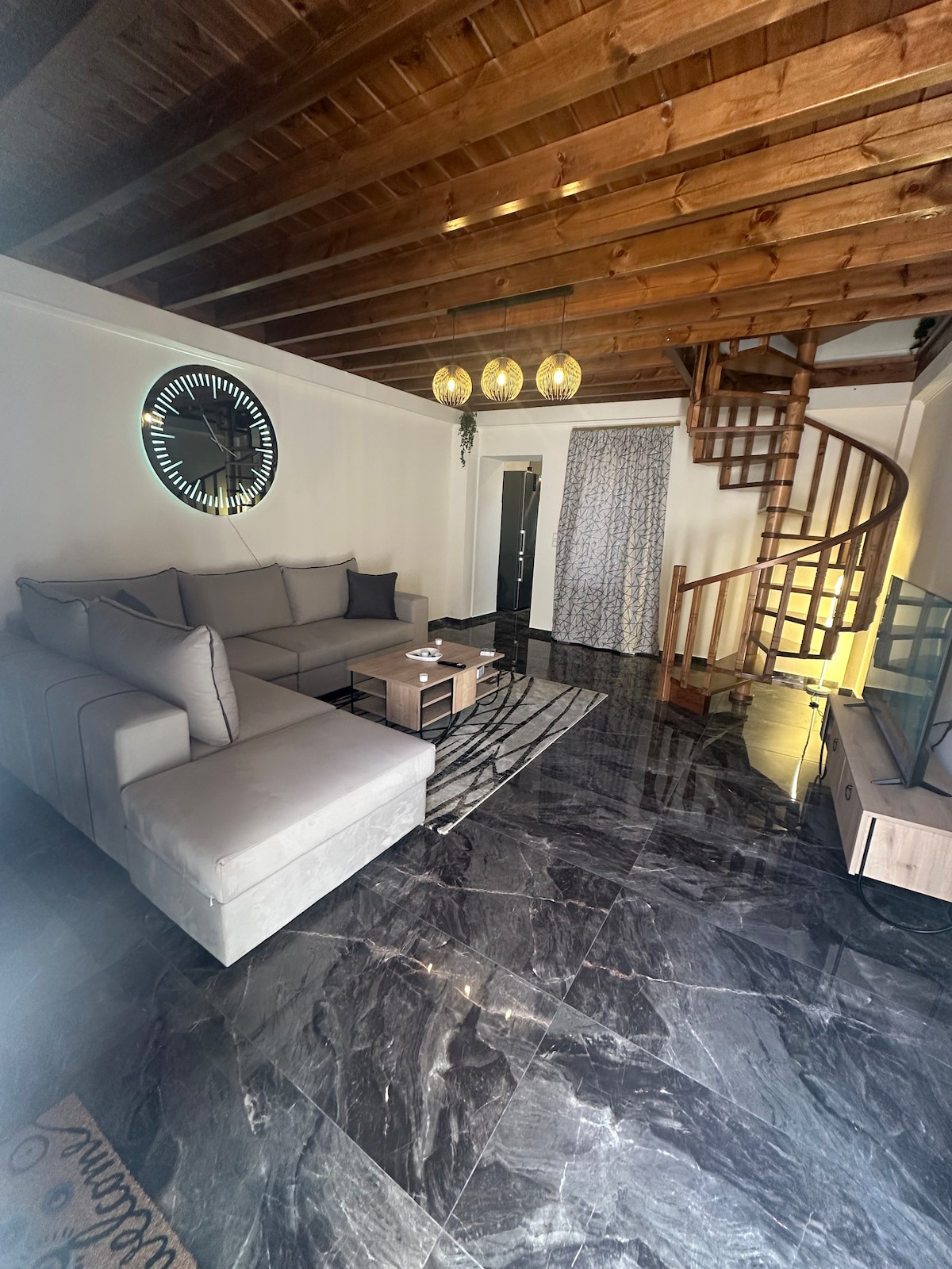 A spacious living area features a large, gray sectional sofa and a glass coffee table. Dark marble flooring reflects the light from the staircase, which spirals upward. A wall clock is mounted prominently, and sheer curtains cover a window, allowing soft light to enter the room.