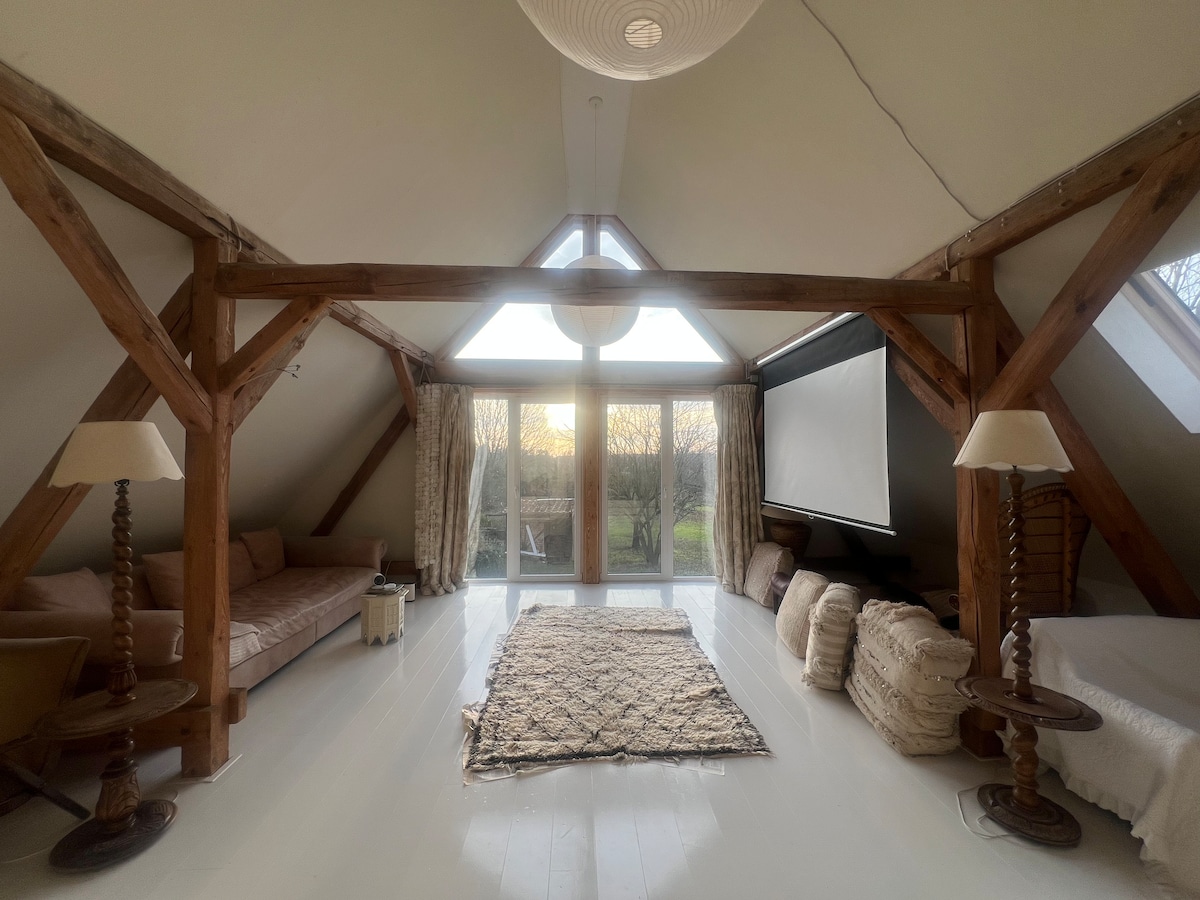 A spacious loft area is highlighted by wooden beams and large glass doors that open to scenic views. A neutral-toned rug sits on light flooring, complemented by a cozy seating arrangement and a retractable screen for entertainment.