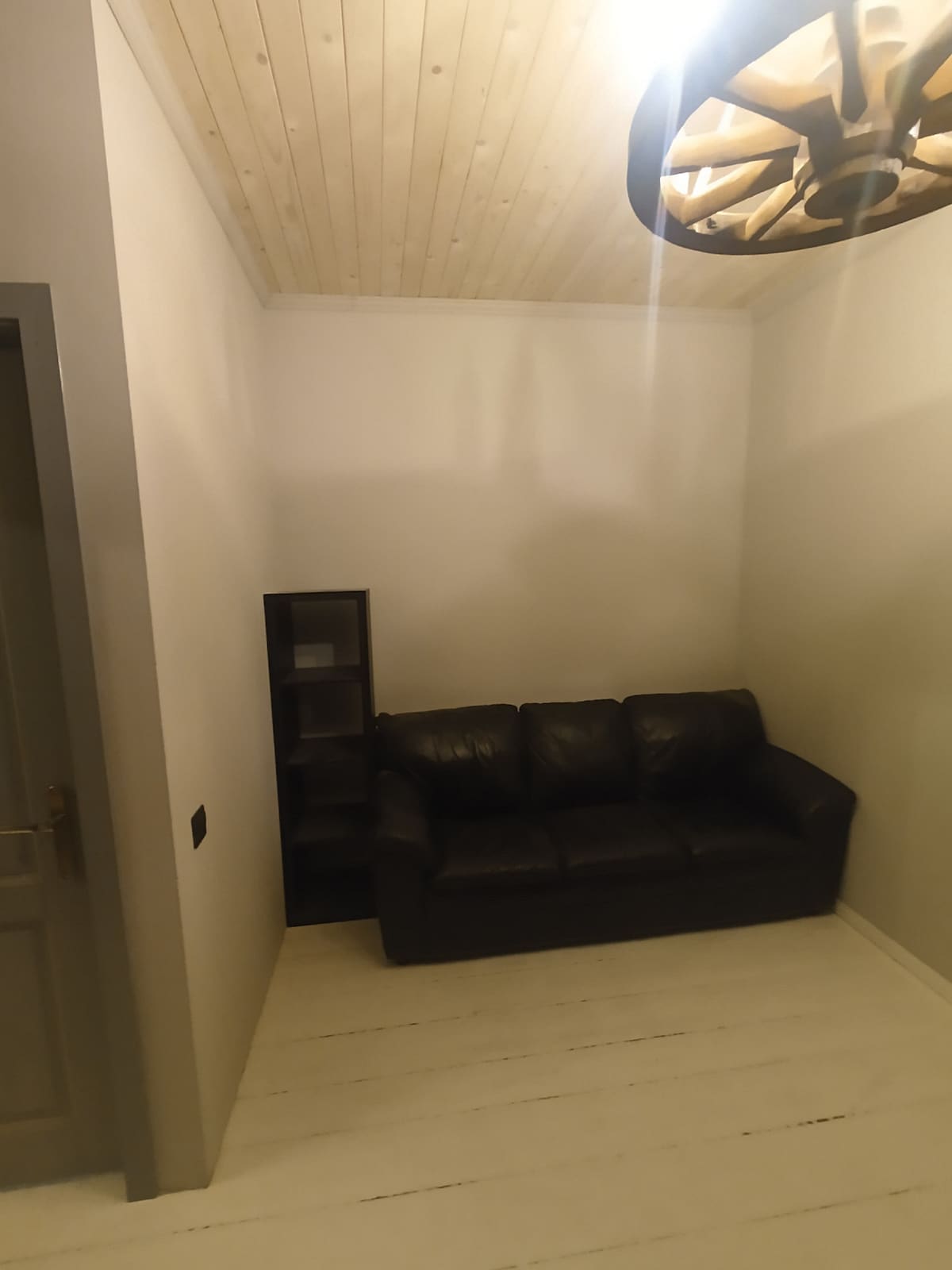 A cozy seating area is featured, showcasing a dark leather sofa against a light-colored wall. A simple bookshelf stands nearby, providing space for storage or display. The ceiling boasts a wooden finish, and a light fixture adds a touch of illumination.