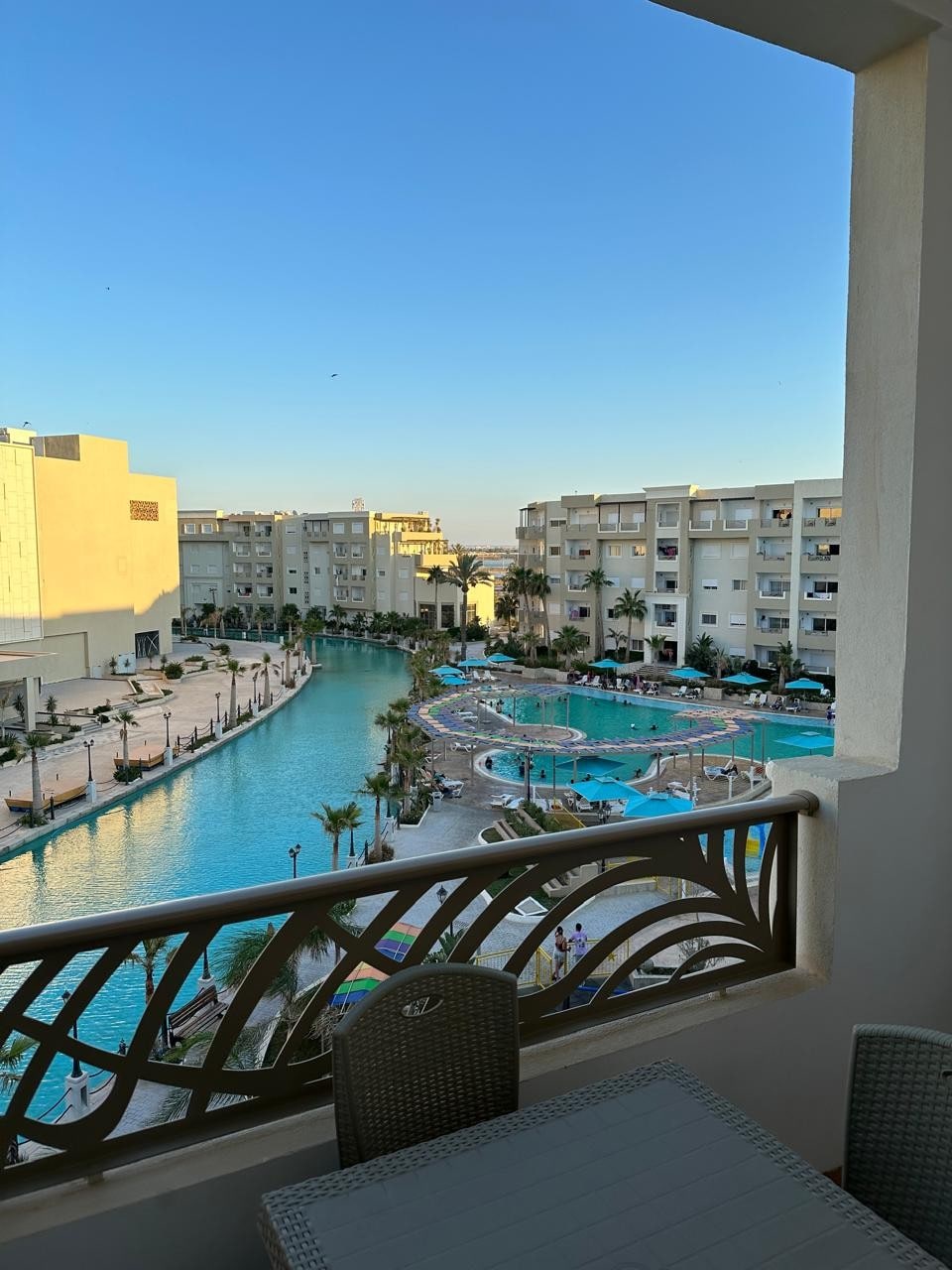 The view from the balcony offers a glimpse of the expansive pool area, featuring several swimming pools with winding paths and lush greenery. Modern buildings are visible on either side, and the blue sky overhead creates a serene atmosphere.