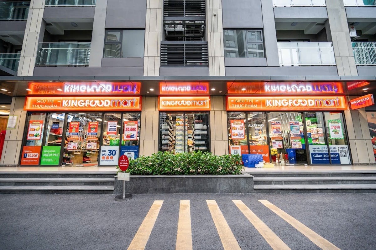 The image displays the exterior of a grocery store named King Food Mart, featuring brightly lit signage. Shelves stocked with various products are visible through the glass entrance. A landscaped area with green plants lines the front, while crosswalk markings are seen on the pavement.