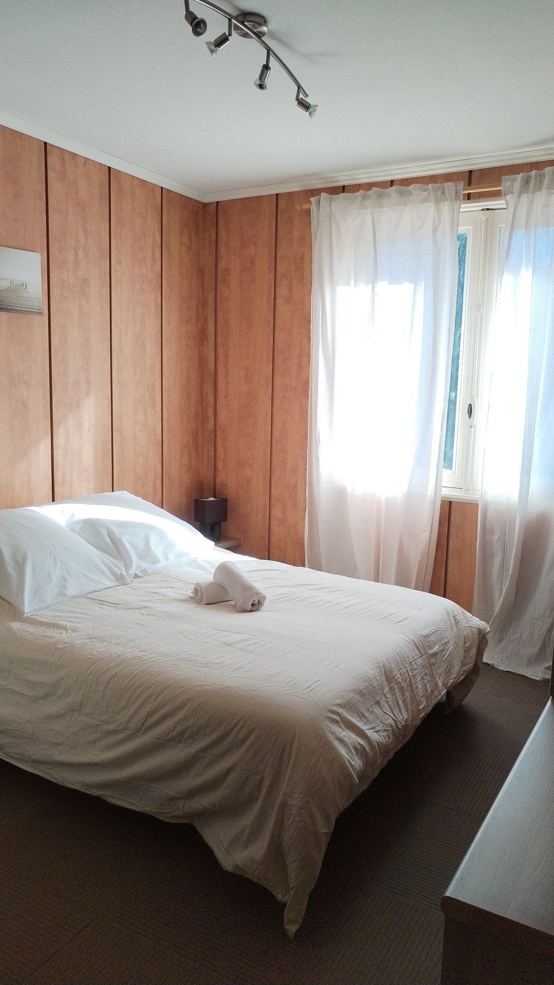 A private room features warm wooden paneling and a spacious bed topped with fresh linens. Natural light filters through a window adorned with sheer curtains. A bedside table with a lamp adds functionality to the cozy atmosphere.