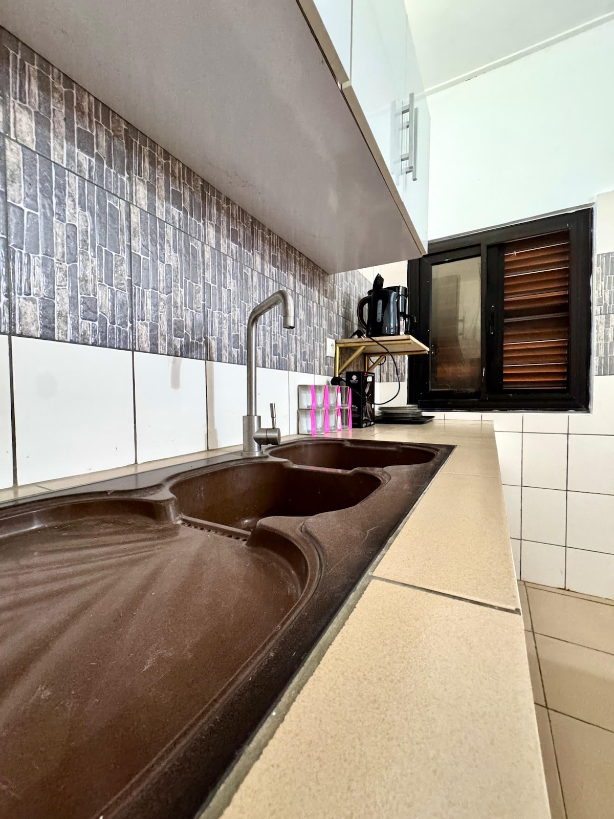 A simple kitchen area features a double sink set against a patterned backsplash. Light-colored countertops hold a coffee maker and utensils. A window with wooden shutters allows natural light to enter, providing a view of the outside.