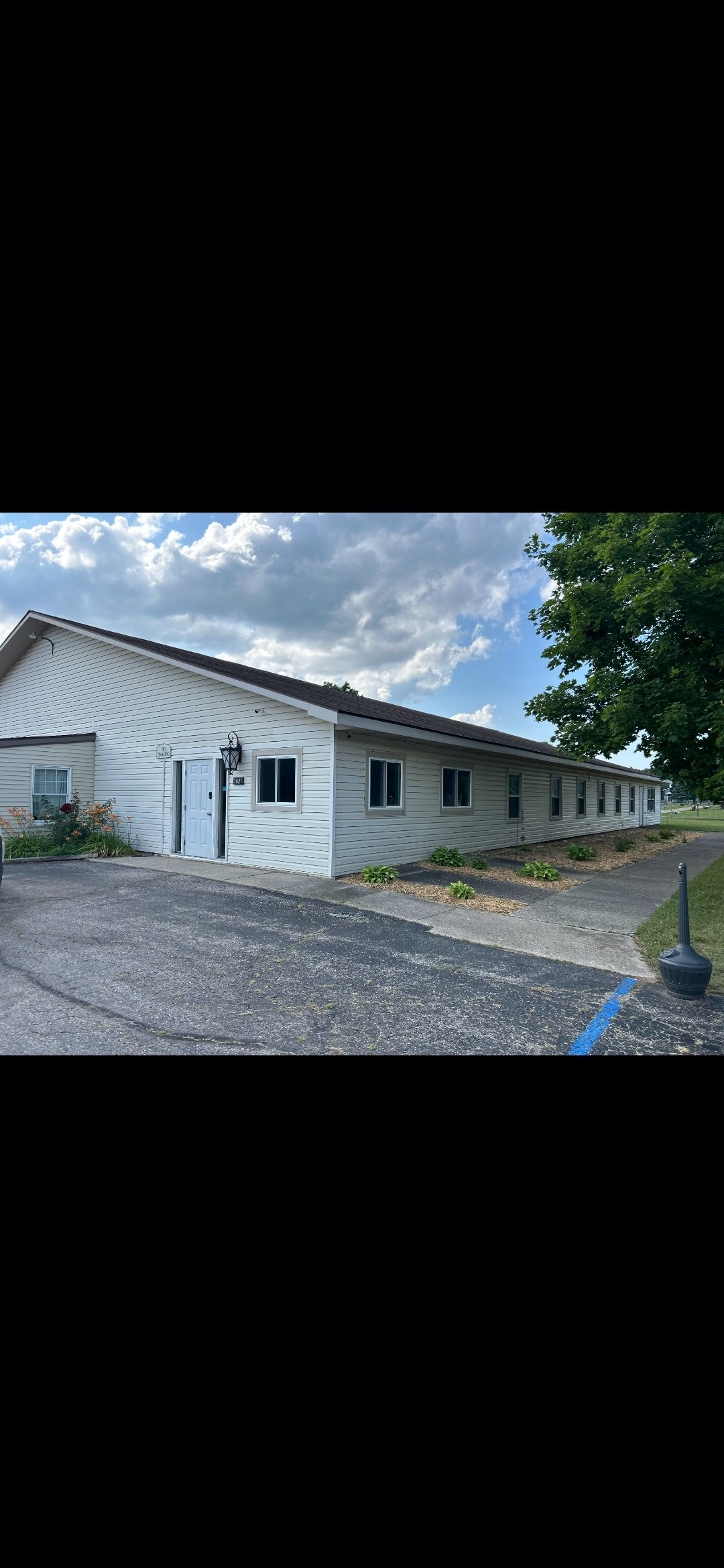 A spacious, single-story retreat with a white exterior is shown. The building features large windows, a welcoming entrance, and a well-maintained path leading to the door. Green shrubbery is present around the property, and a blue parking line is visible beside the building.