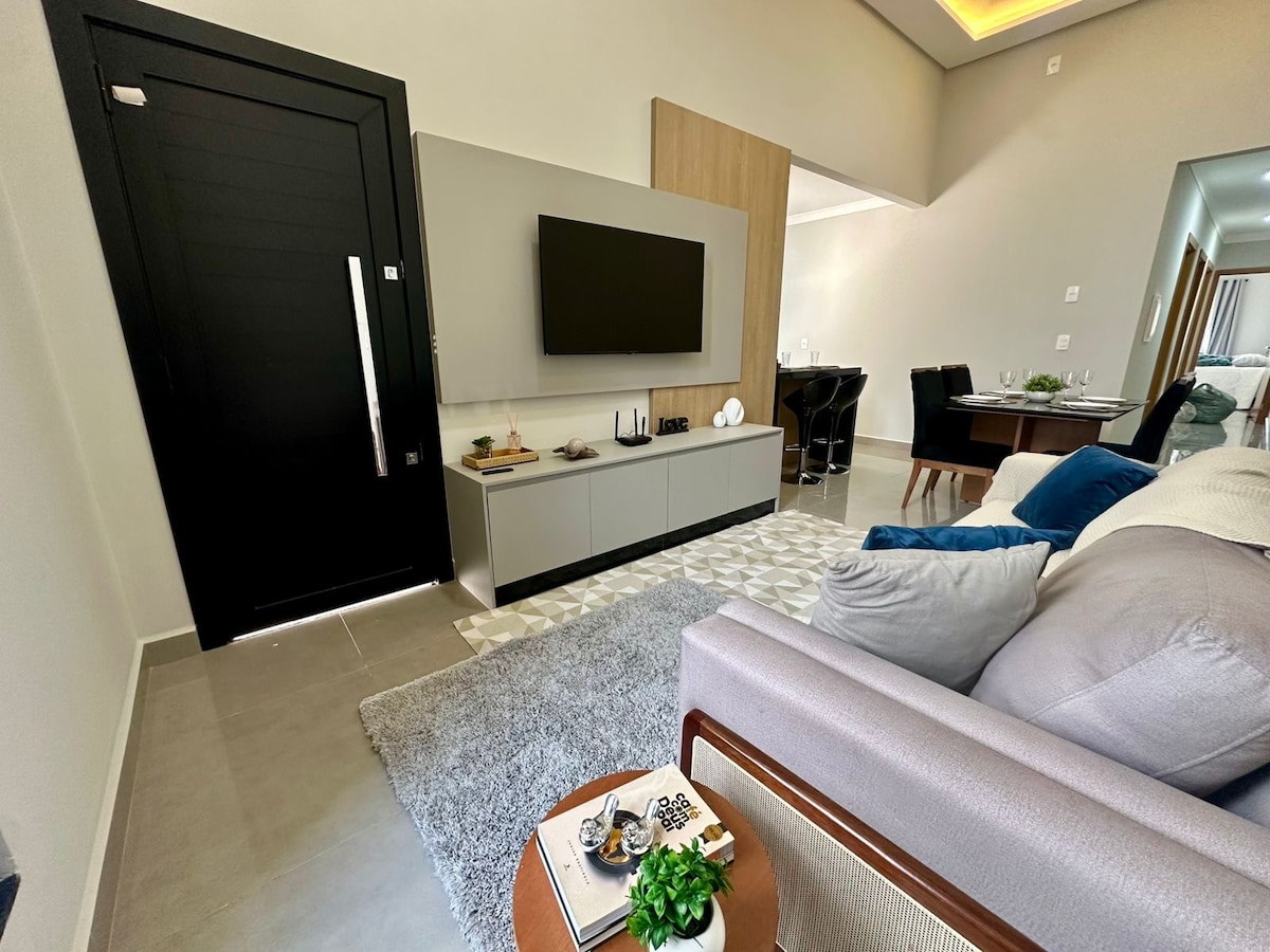 A modern living area is presented with a sleek television mounted on a light grey wall unit. A soft area rug covers the floor, complementing the neutral tones of the furnishings. A cozy sofa is positioned in the foreground, featuring decorative pillows and a small table with greenery.