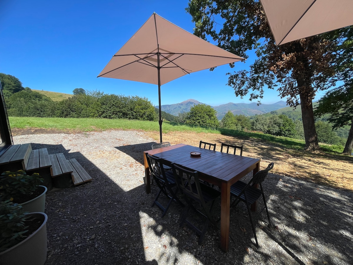 An outdoor dining area is shown under large umbrellas, offering shade. A wooden table with several chairs is positioned on gravel, surrounded by greenery and views of rolling mountains in the background. The bright sky contributes to a serene atmosphere.